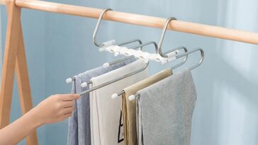 an image of a stainless steel multi-layer pants hanger. It should be hanging in a modern wardrobe filled with various types of garments like pants, scarves, and towels. The hanger should showcase a sleek, space-saving design with up to five horizontal rods, each holding folded clothing. Include a rotating hook at the top, and highlight the detachable rods for easy access. The background should be a neatly organized wardrobe with a minimalist, bright, and clutter-free aesthetic."